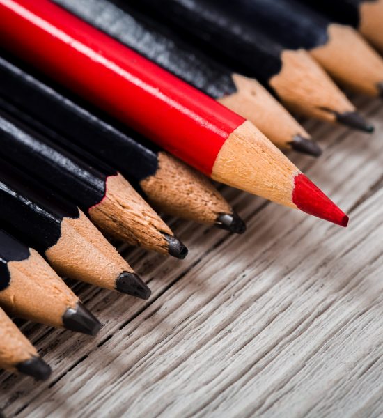 Red pencil stands out from the crowd of black pencil on a wooden white background. Leadership, uniqueness, independence, initiative, strategy, disagree, think, different, concept of business success